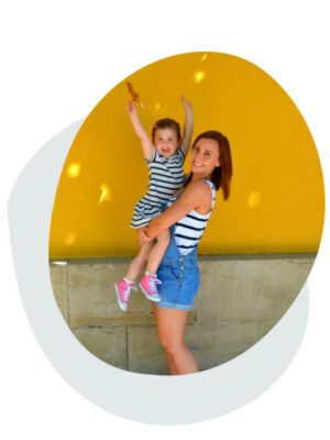 mother wearing dungarees with daughter in arms in front of yellow wall