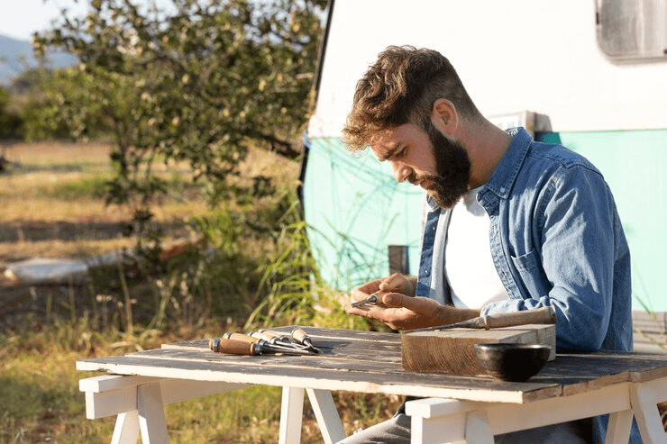 man sitting at picnic bench outside using tools with caravan behind him