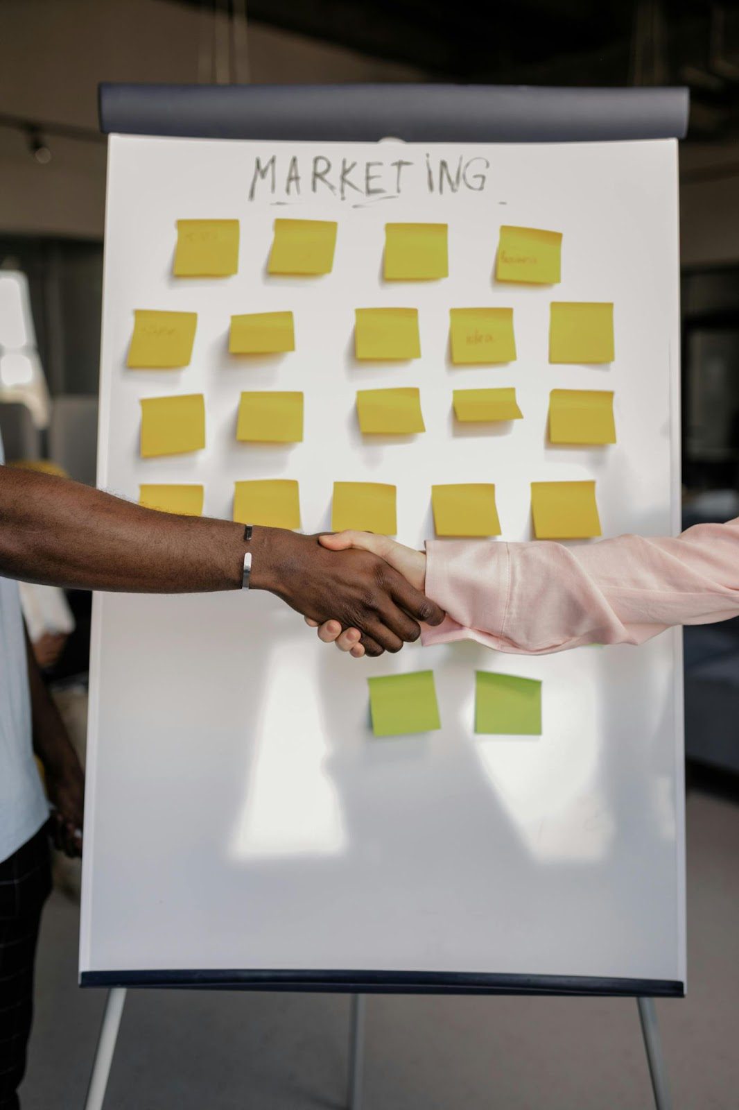 board with 'marketing' written at the top and yellow sticky notes all over it, with two people shaking hands in front of it