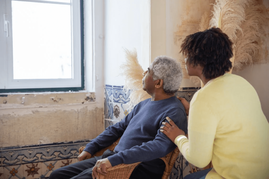 man sitting in chair looking out window with women crouching down behind him