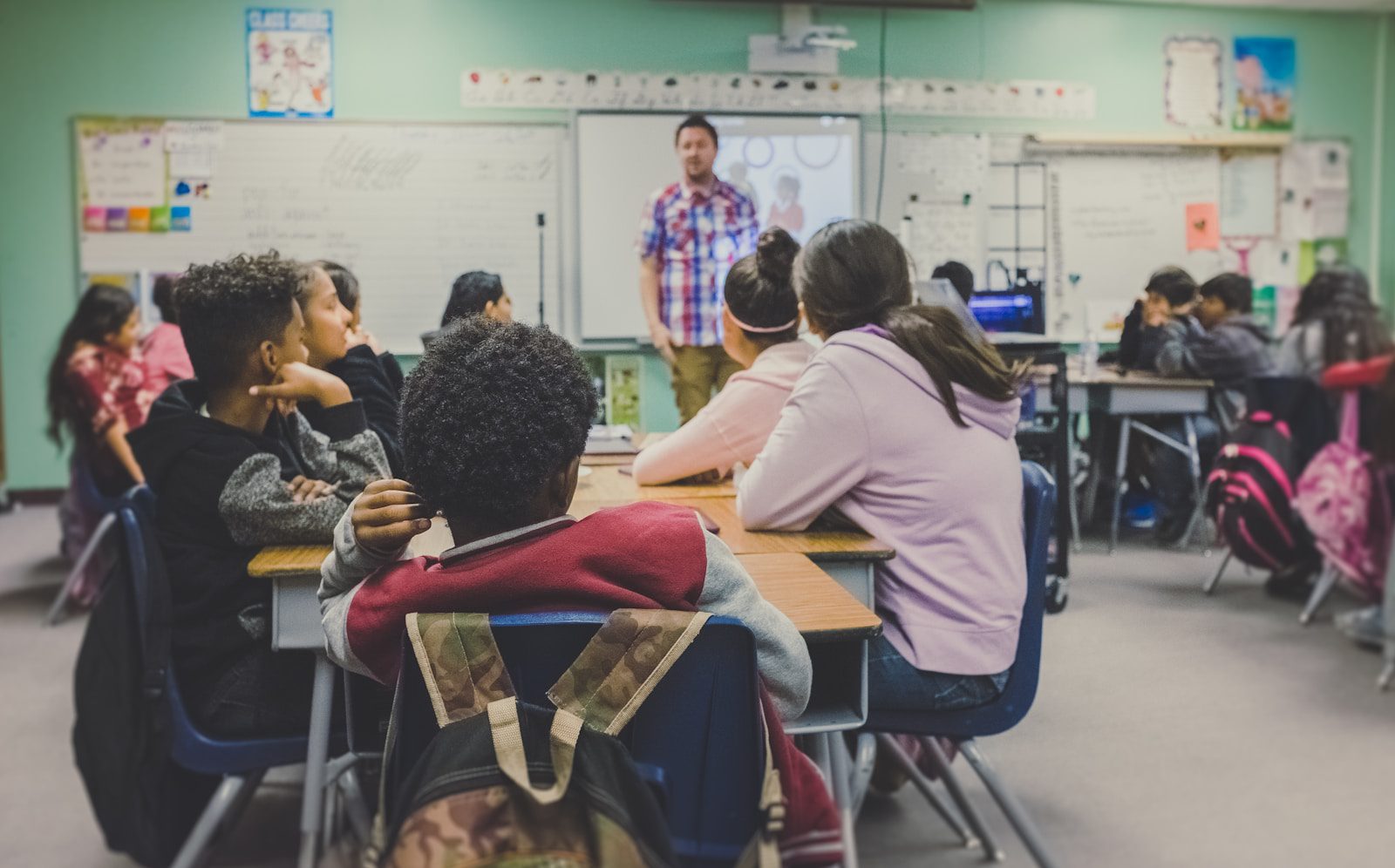 male teacher standing at front of class with children sitting on chairs at tables