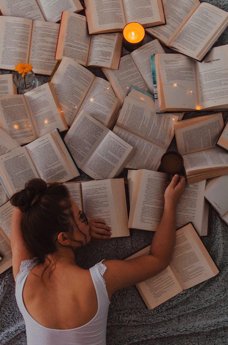 woman in white tank top with upper body and face on piles of open books, a mug of tea in her hand and a candle